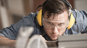 Arbeit - Das Bild zeigt einen Mann mit Ohrenschützer und Schutzbrille. Er steht vor einer Werkbank.